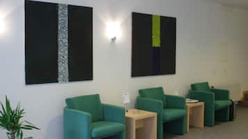 Un área de espera con sillas verdes, una mesa pequeña y dos cuadros abstractos en la pared.
