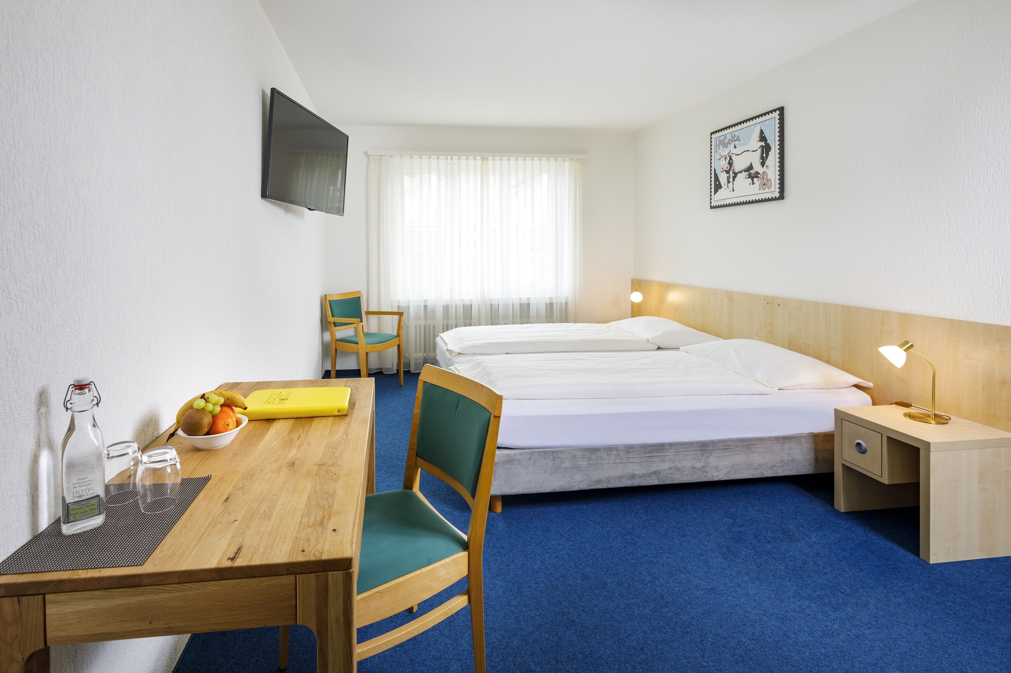 A hotel room with a bed, a desk with a chair, a small table with a bowl of fruit, a bedside lamp, and a TV on the wall.