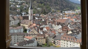 Blick durch einen hölzernen Bogen auf eine Stadt mit einem markanten Glockenturm, umgeben von Gebäuden und einer Bergkulisse.
