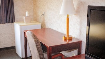 Una habitación de hotel pequeña con una mesa de madera, una lámpara, un televisor, un refrigerador minibar y una silla.
