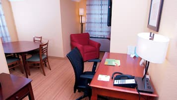 Une chambre d’hôtel avec une table à manger en bois, deux chaises, un fauteuil rouge, un bureau avec une lampe, un téléphone et un téléviseur fixé au mur.