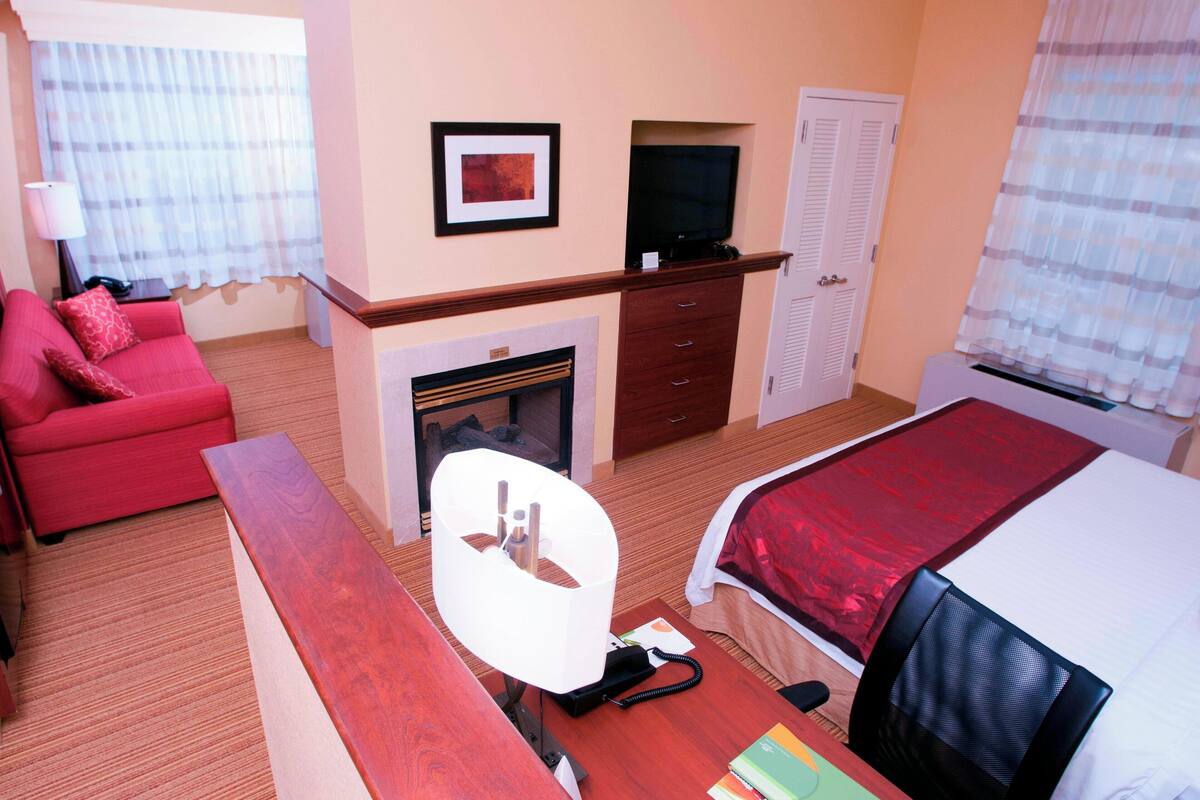 Ein Hotelzimmer mit rotem Sofa, einem Bett mit roter Decke, einem Fernseher auf einem Holzregal, einem Kamin und einem Schreibtisch mit Lampe und Telefon.