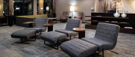 A modern hotel lobby with grey upholstered seating, a coffee table, and a lamp.