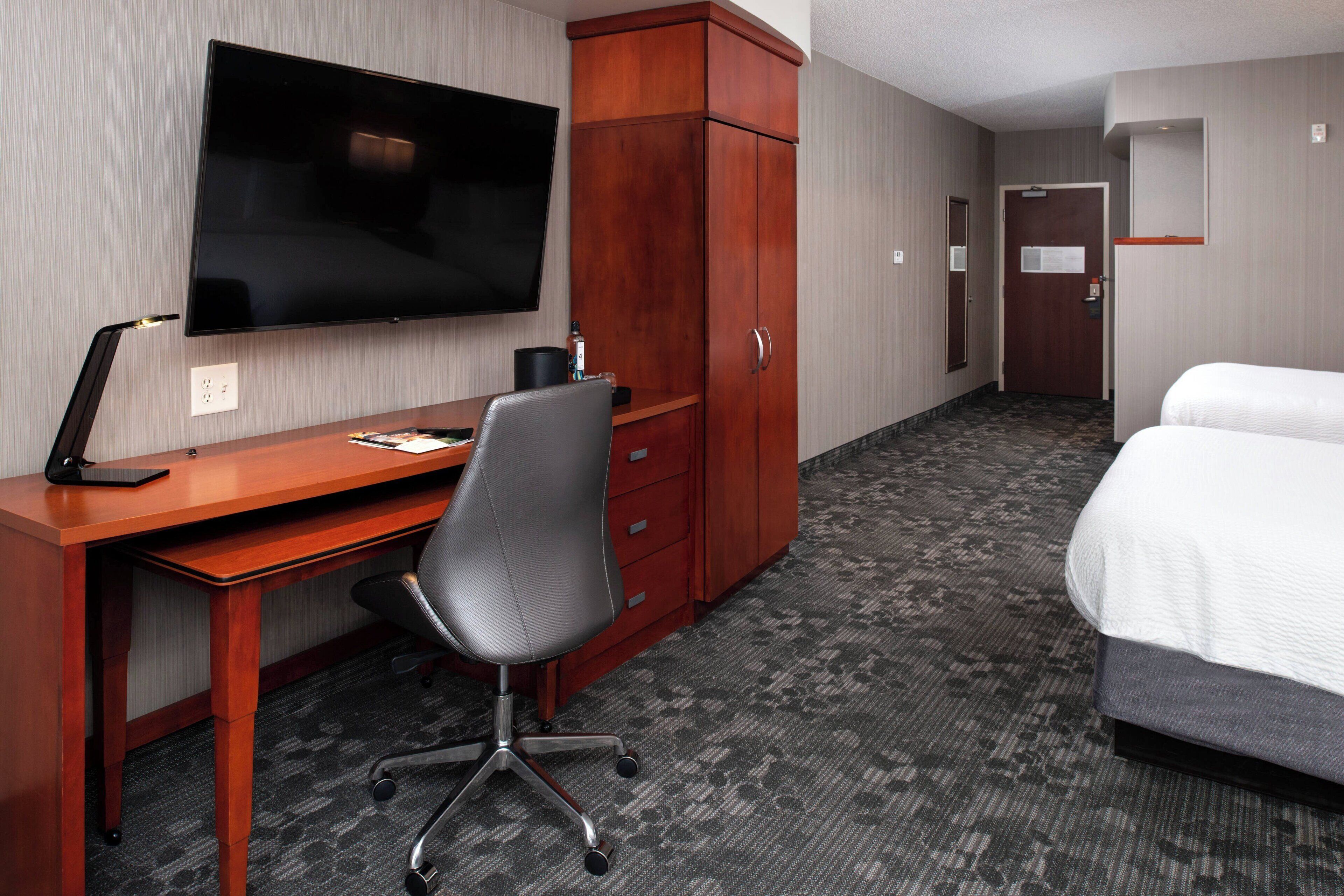 Habitación de hotel con un escritorio de madera, una silla de oficina negra, un televisor de pantalla plana montado en la pared, un armario, una cama con ropa blanca y piso alfombrado.