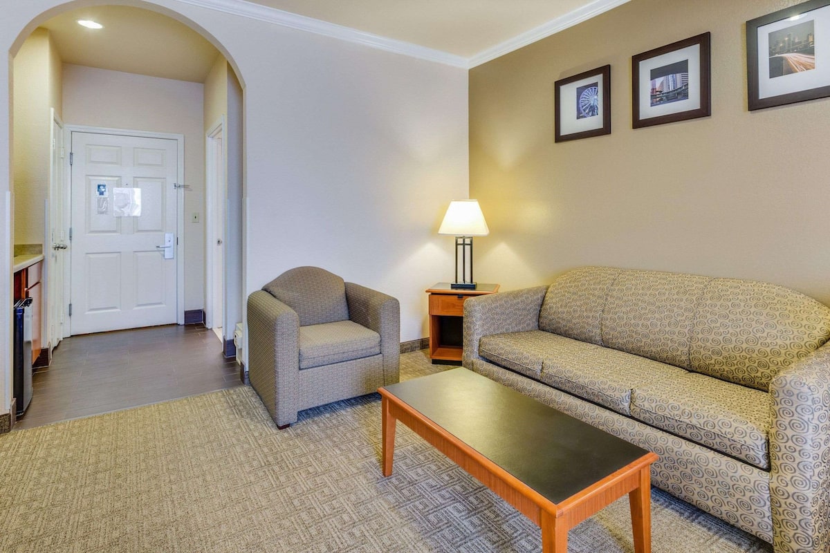 A hotel room with a sofa, armchair, and a small table. There is a door leading to another room and a lamp on a side table.