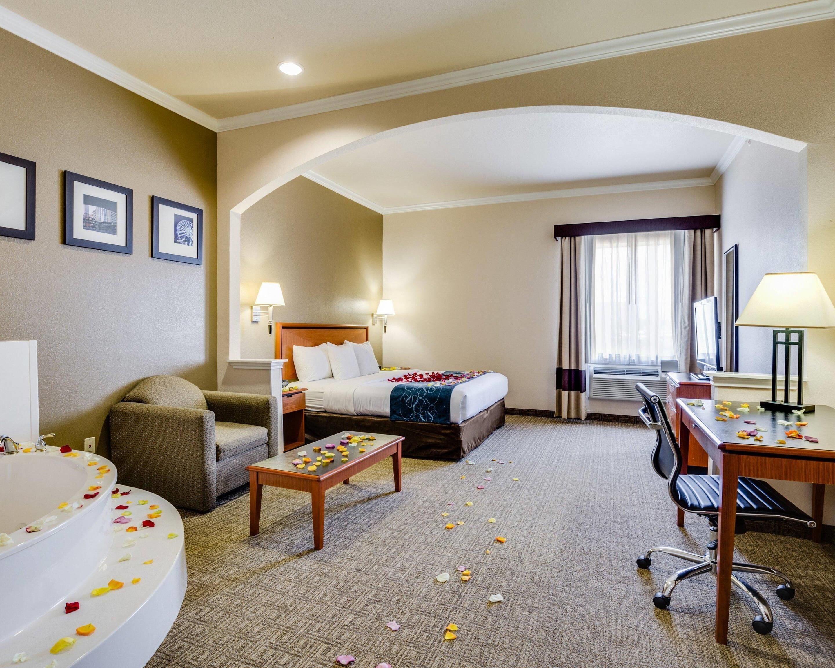 A hotel room with a bed, a desk, a chair, a sofa, a bathroom with a bathtub, and a large window with curtains.