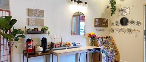 Una cocina con cafetera, un lavamanos y una pequeña mesa de comedor. Hay platos decorativos en la pared y una planta en maceta.