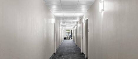 A long, narrow hallway with white walls and a dark patterned floor, leading to a room with a large window.