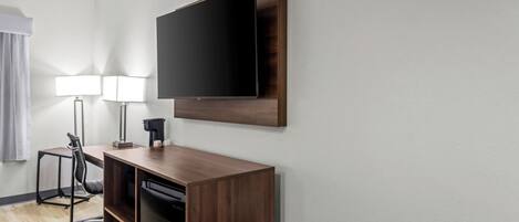 Habitación de hotel con escritorio de madera, televisor de pantalla plana montado en la pared, una silla, una lámpara y una ventana con cortinas.