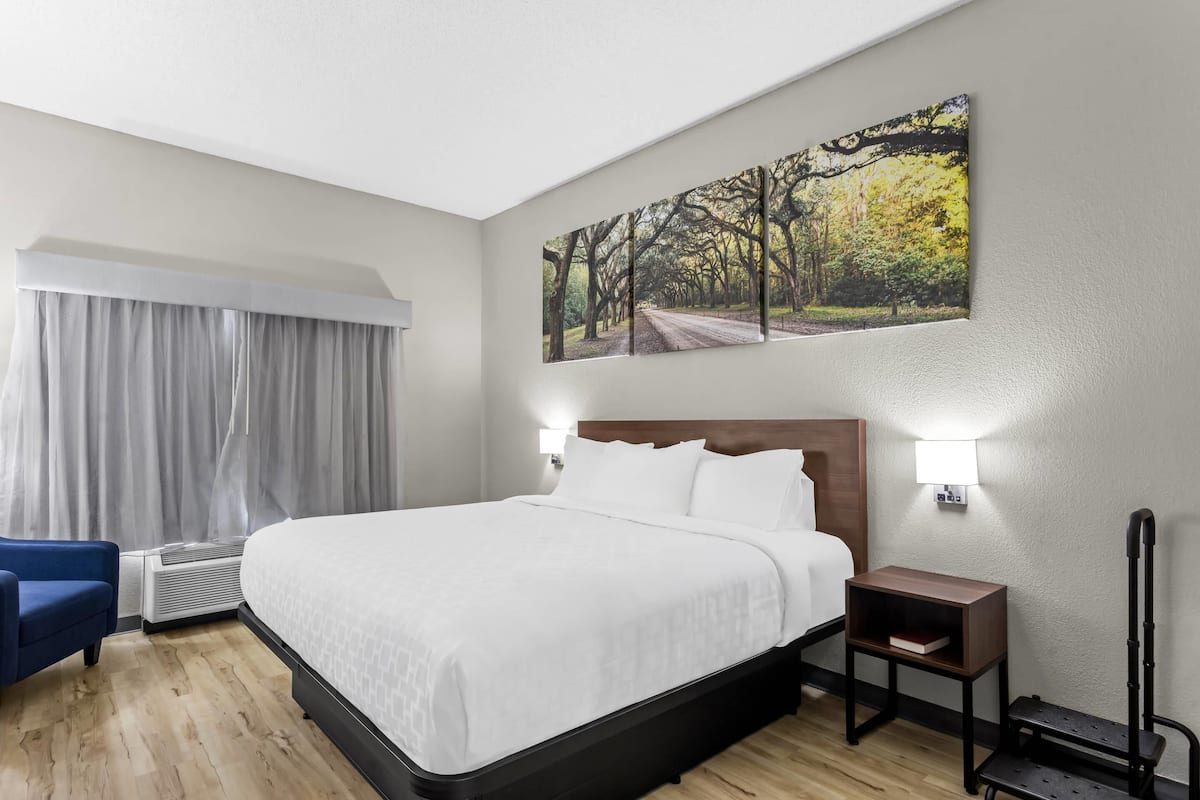 A hotel room with a large bed, a bedside table, a chair, a wall-mounted painting, and a window with curtains.