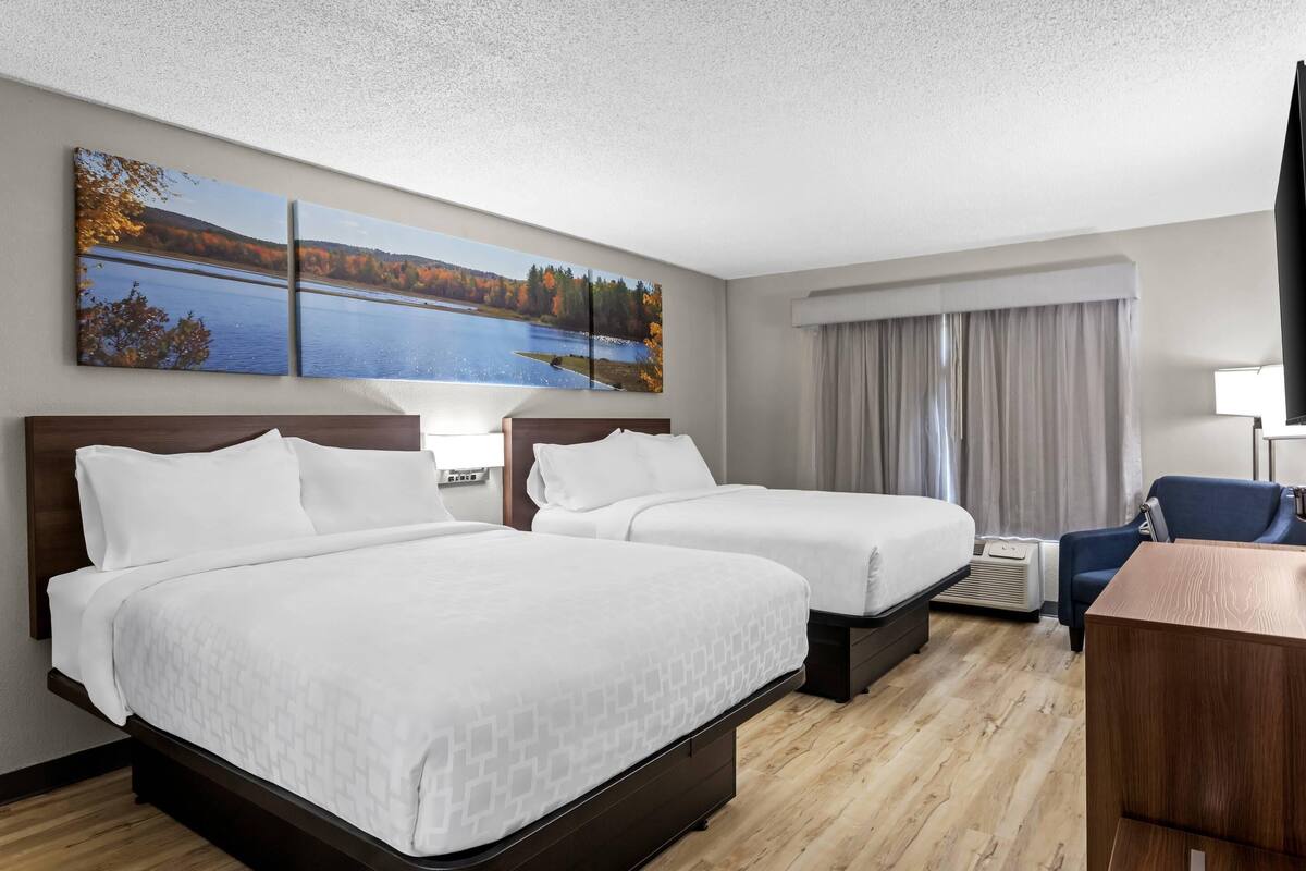 A hotel room with two beds, a desk, a chair, a large window with curtains, and a painting of a lake.