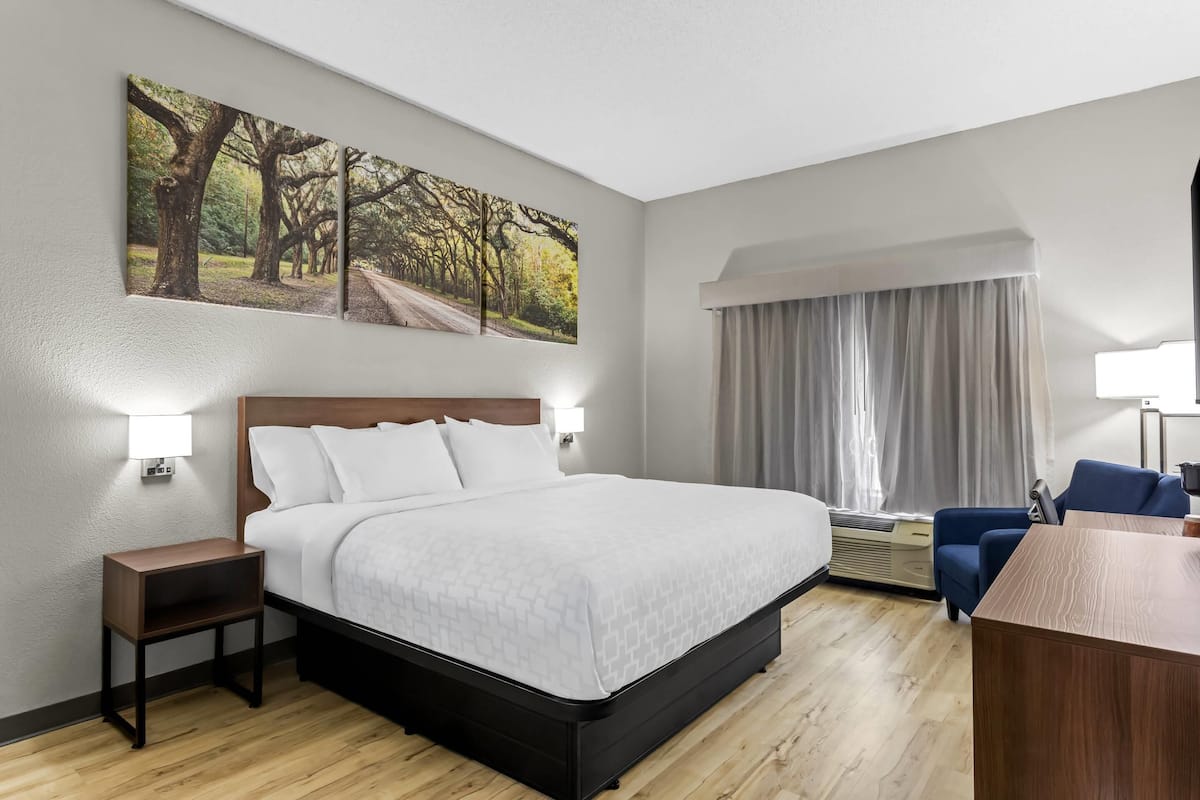 A hotel room with a bed, bedside lamps, a desk, a chair, a painting on the wall, and a window with curtains.
