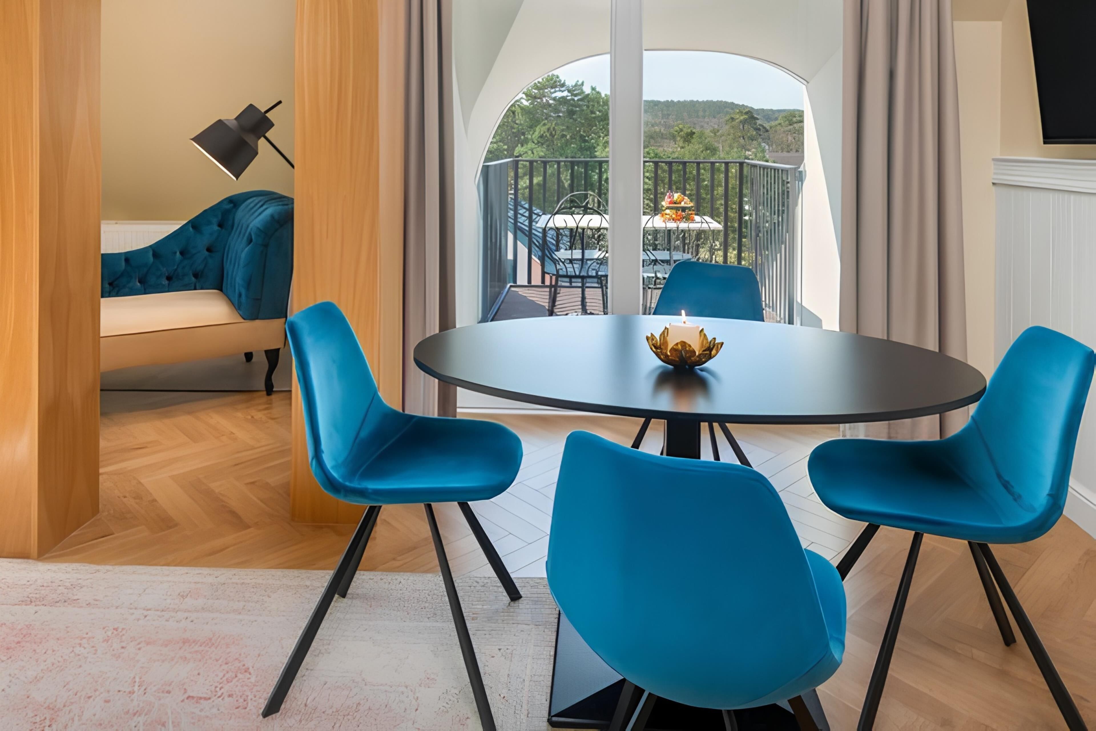 Ein modernes Zimmer mit einem runden Tisch, vier blauen Stühlen, einer Couch und einem Balkon mit Gartenmöbeln.