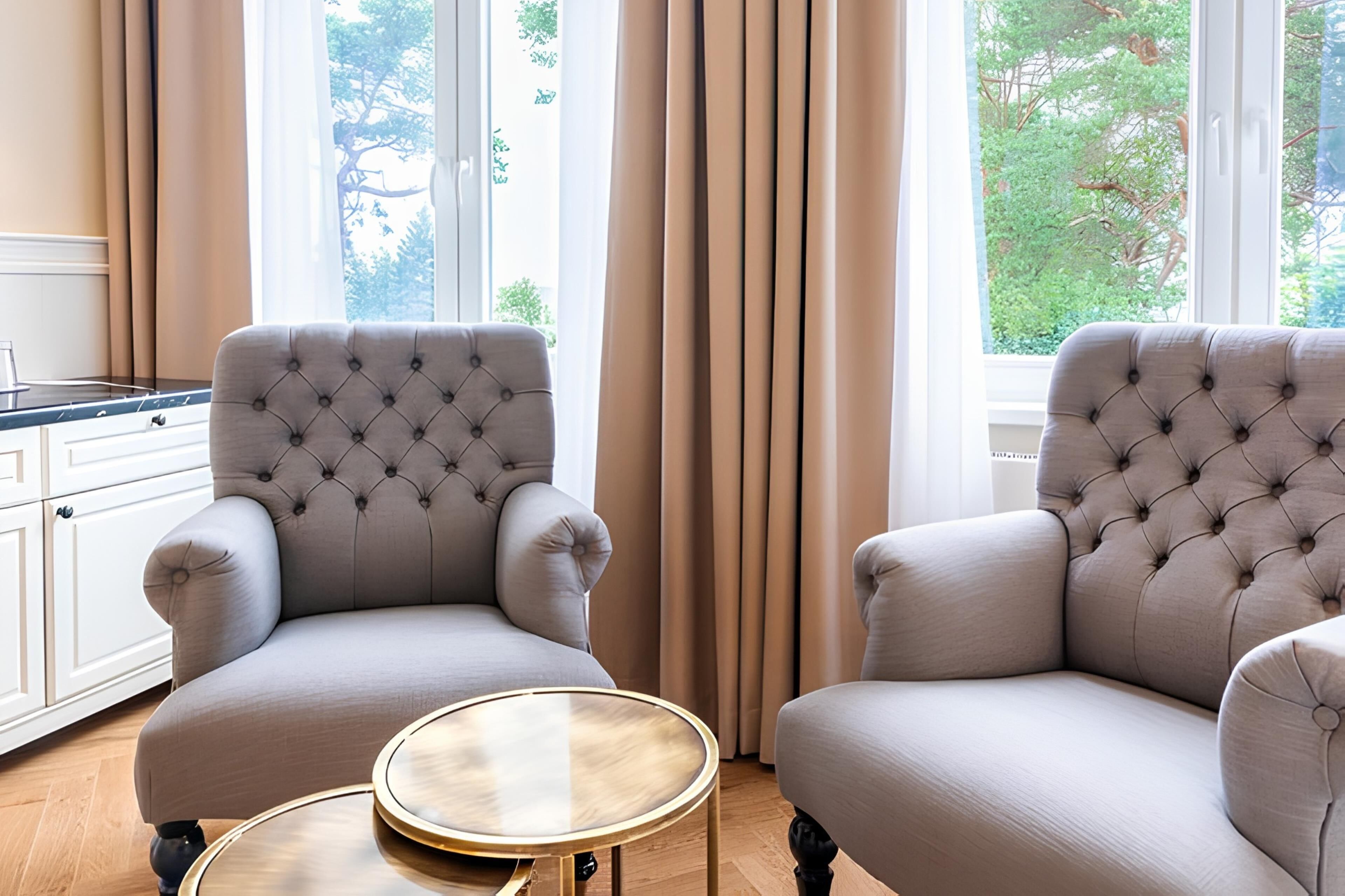 Ein Zimmer mit zwei gepolsterten Sesseln, einem runden Glastisch und Blick auf Grünanlagen durch große Fenster.