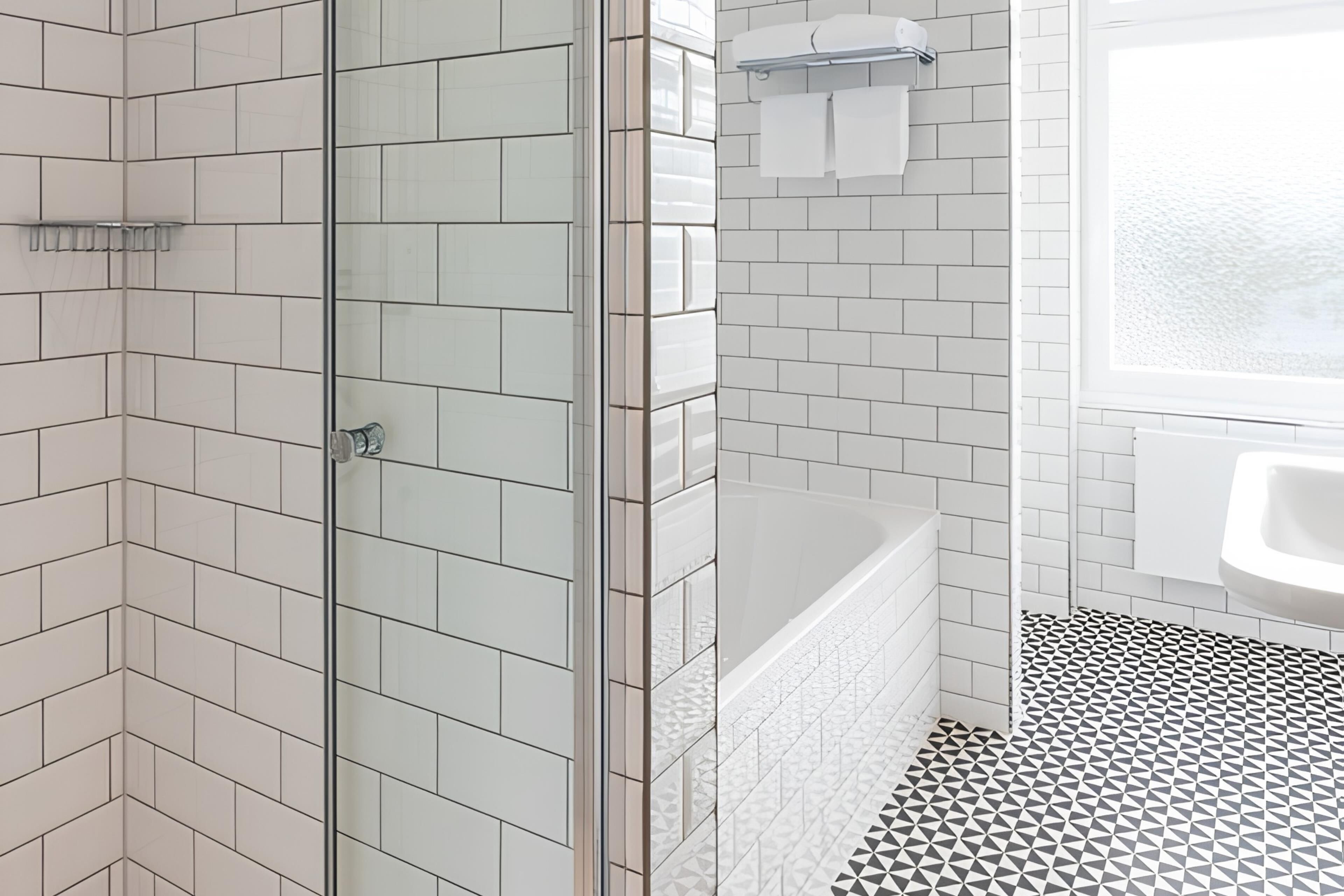 Un baño moderno con azulejos blancos tipo metro, cabina de ducha de vidrio, bañera y suelo con estampado geométrico.