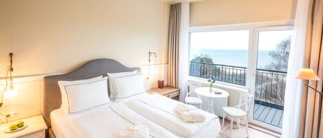 A hotel room with a large bed, a bedside table with a lamp, a small table with chairs, and a balcony with a view of the sea.