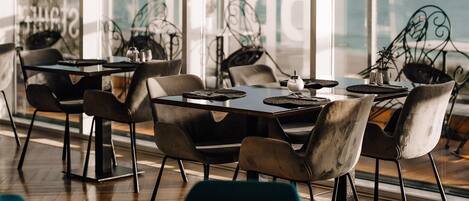 A restaurant with tables and chairs, large windows, and a view of the sea.