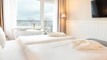 A hotel room with a bed, a small table with a vase, a chair, a TV mounted on the wall, and a balcony with a railing.