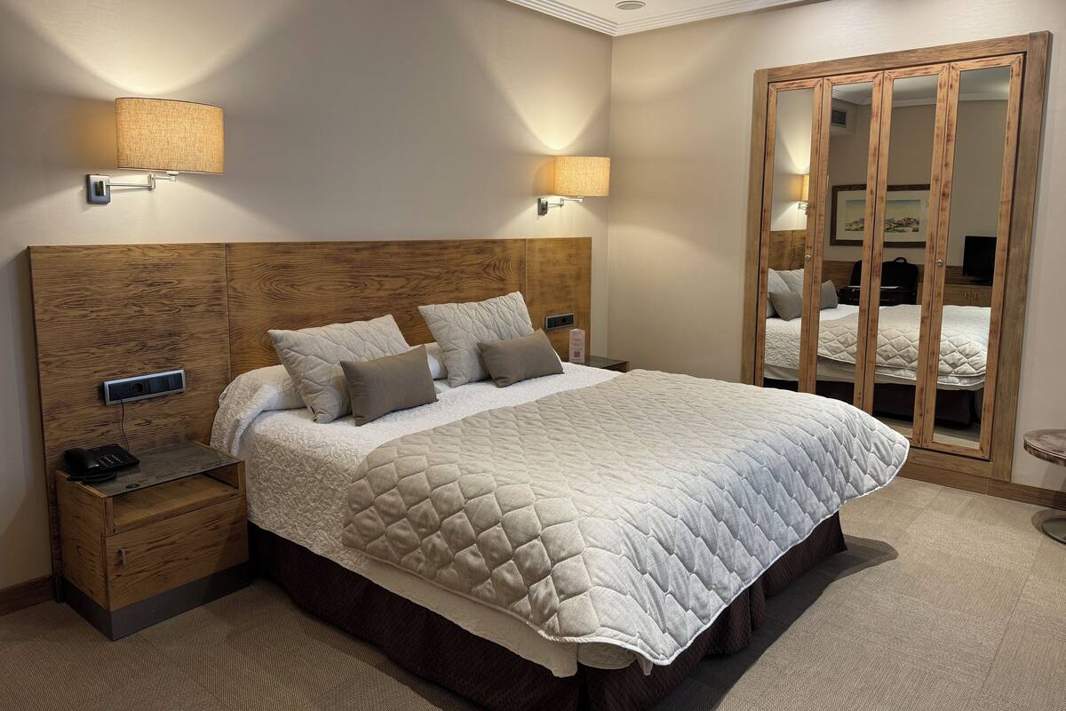 A hotel room with a wooden headboard, a large bed with a quilted duvet, bedside lamps, a nightstand with a phone, and a wardrobe with a mirror.
