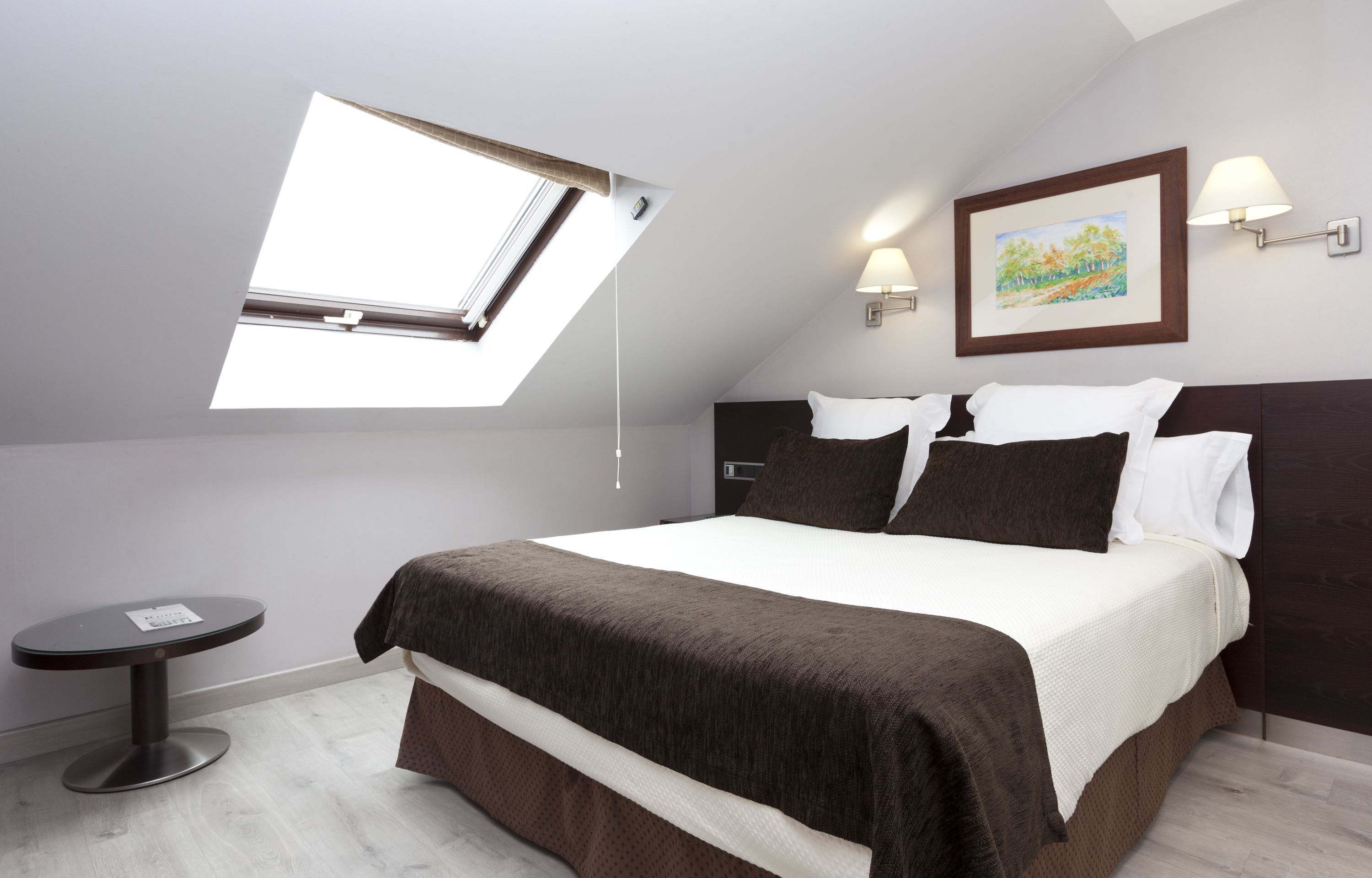 Un dormitorio moderno con tragaluz, cama con ropa de cama blanca y marrón, una mesita redonda y un cuadro enmarcado en la pared.