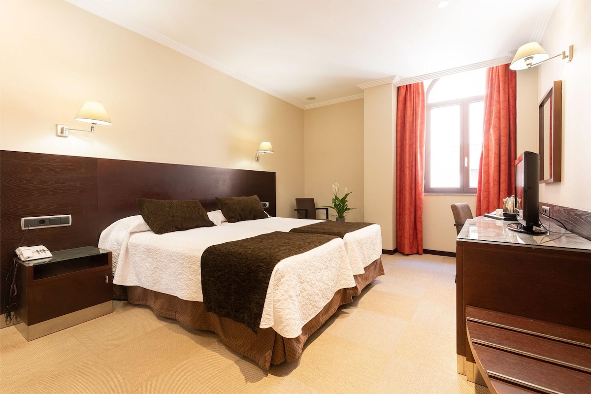A hotel room with a large bed, bedside tables, a desk with a computer, and a window with curtains.