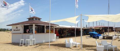 A beachside bar with a Heineken sign, white chairs, and a canopy.