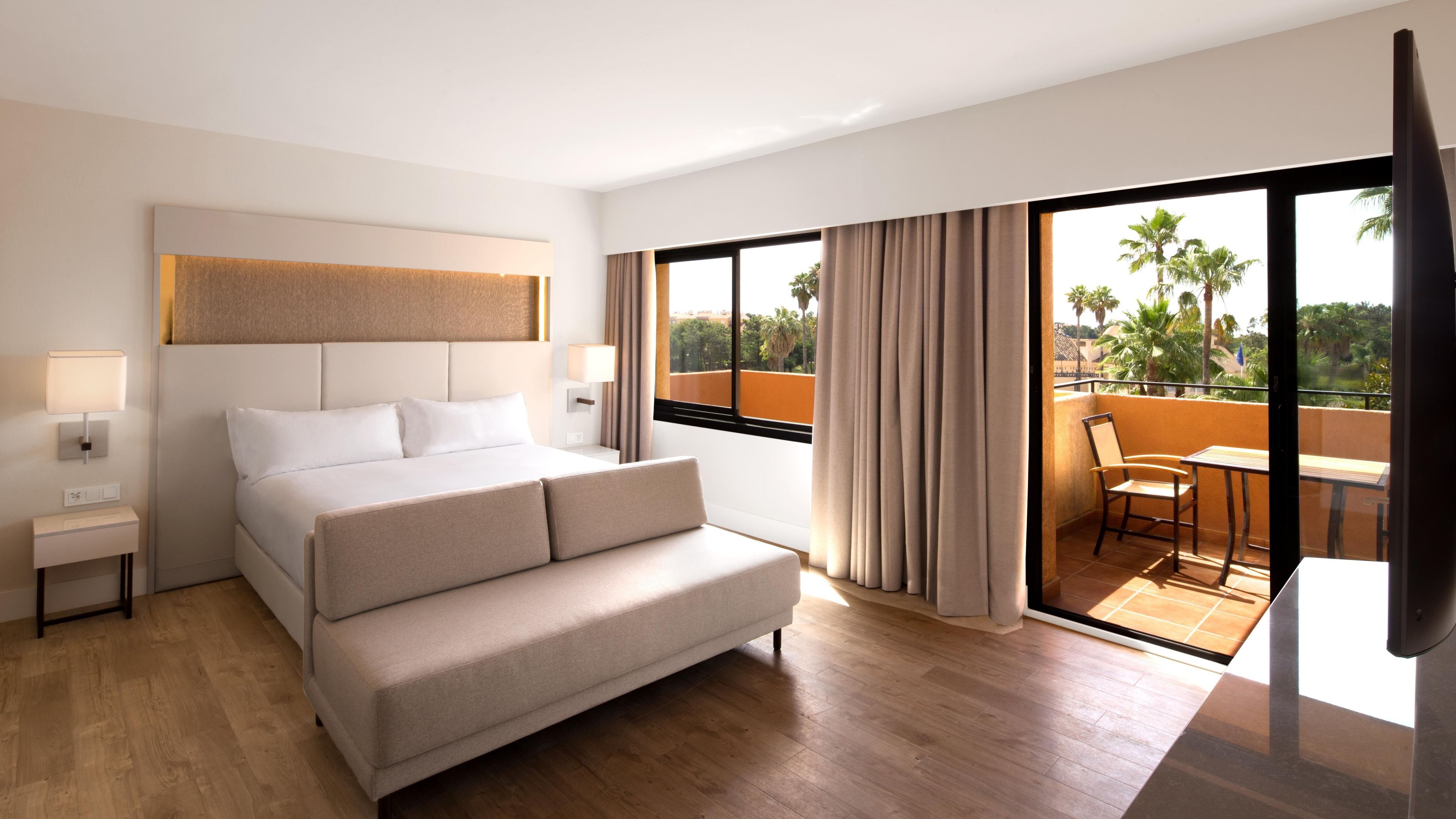 Una habitación de hotel con cama, mesitas de noche, televisor, balcón con mesa y sillas, y vistas a palmeras.