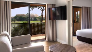A modern hotel room with a large sofa, a round wooden table, a flat-screen TV, and a balcony with a view of the outdoors. - DoubleTree by Hilton Islantilla Beach Golf Resort (Lepe)