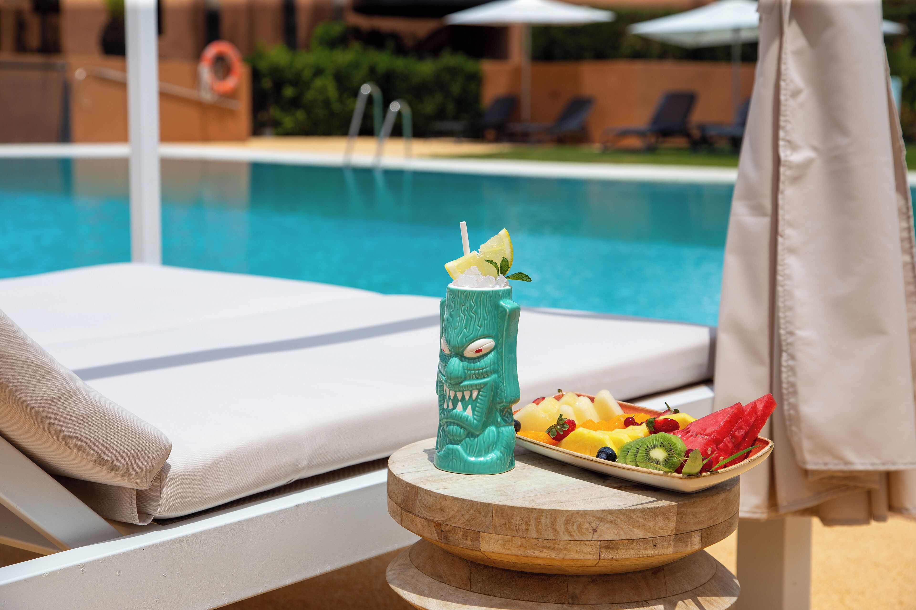 A poolside setting with a white lounge chair, a wooden table with a drink and fruit, and a clear view of the pool.