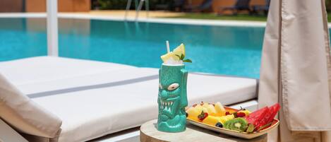 A poolside setting with a white lounge chair, a wooden table with a drink and fruit, and a clear view of the pool.