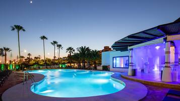 Piscina con sillones de descanso y palmeras, rodeada por un edificio con zona cubierta.