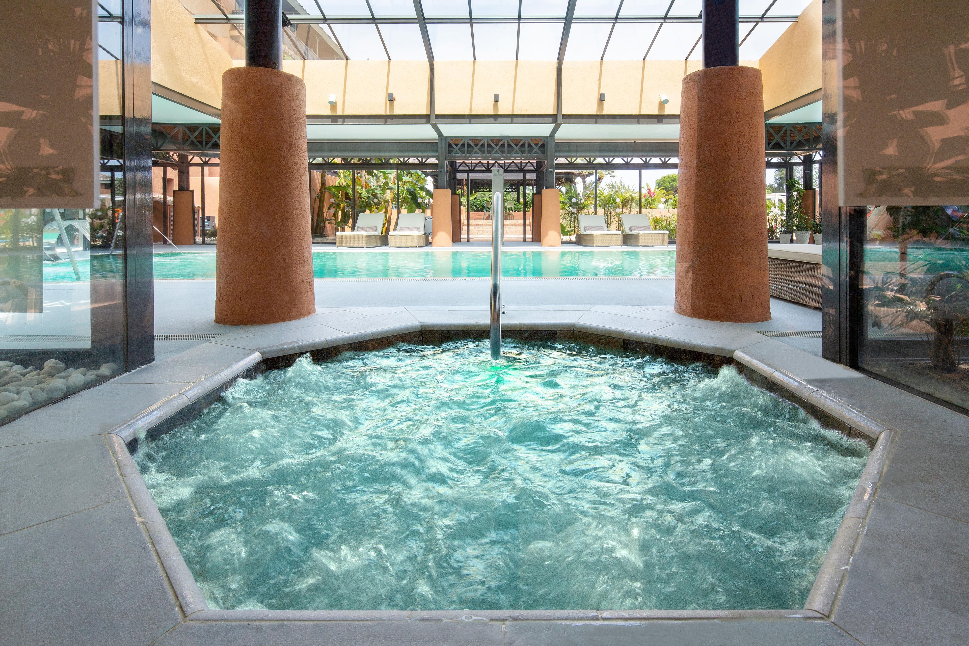 Un área de hidromasaje con piscina al fondo, rodeada de sillas de descanso y un paseo cubierto.