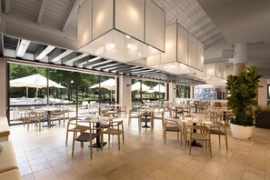 A spacious indoor dining area with multiple tables and chairs, large windows offering views of the outdoors, and a ceiling with geometric light fixtures. - DoubleTree by Hilton Islantilla Beach Golf Resort (Lepe)