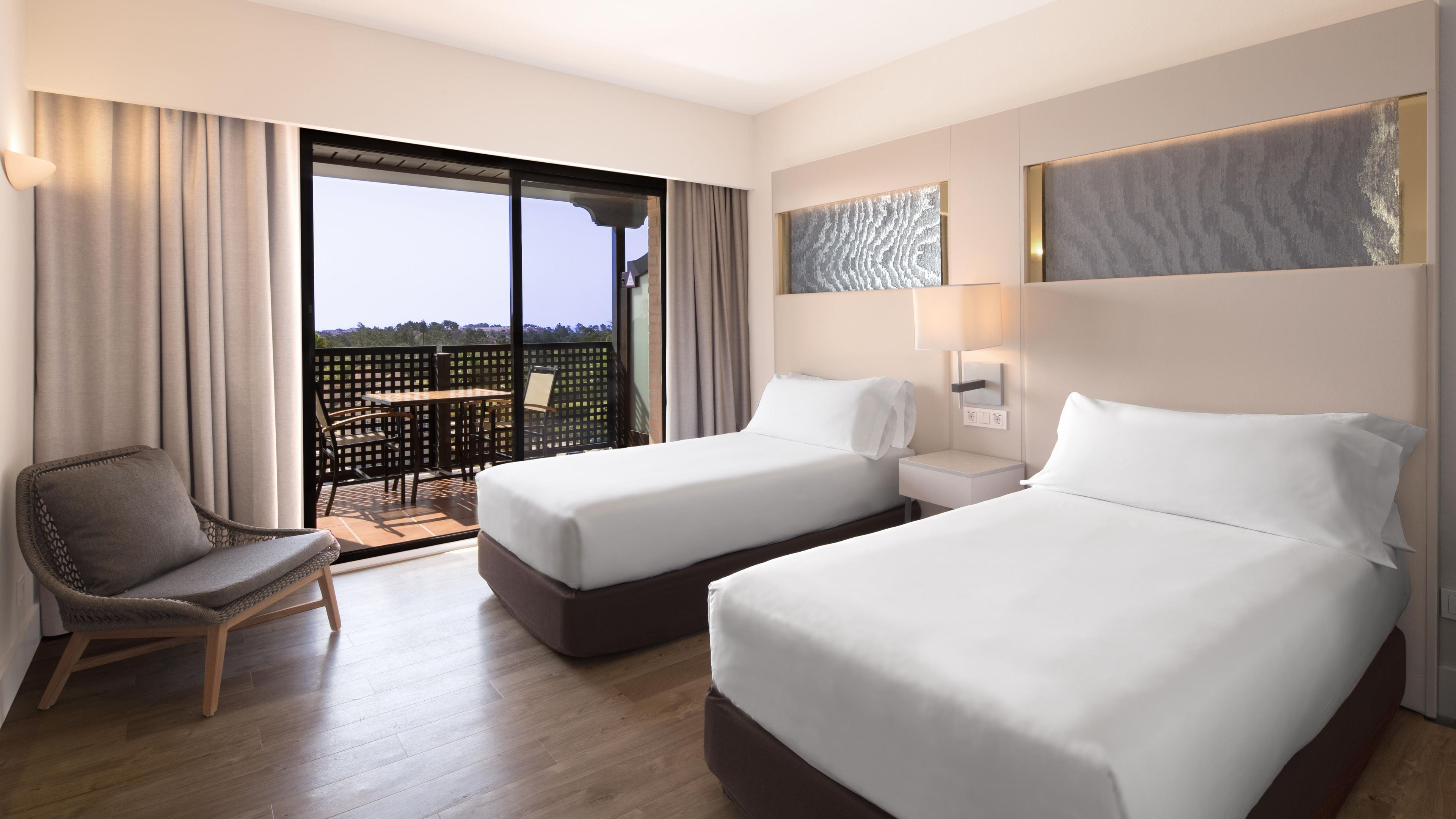 A hotel room with two beds, a chair, a table, and a balcony with a view.