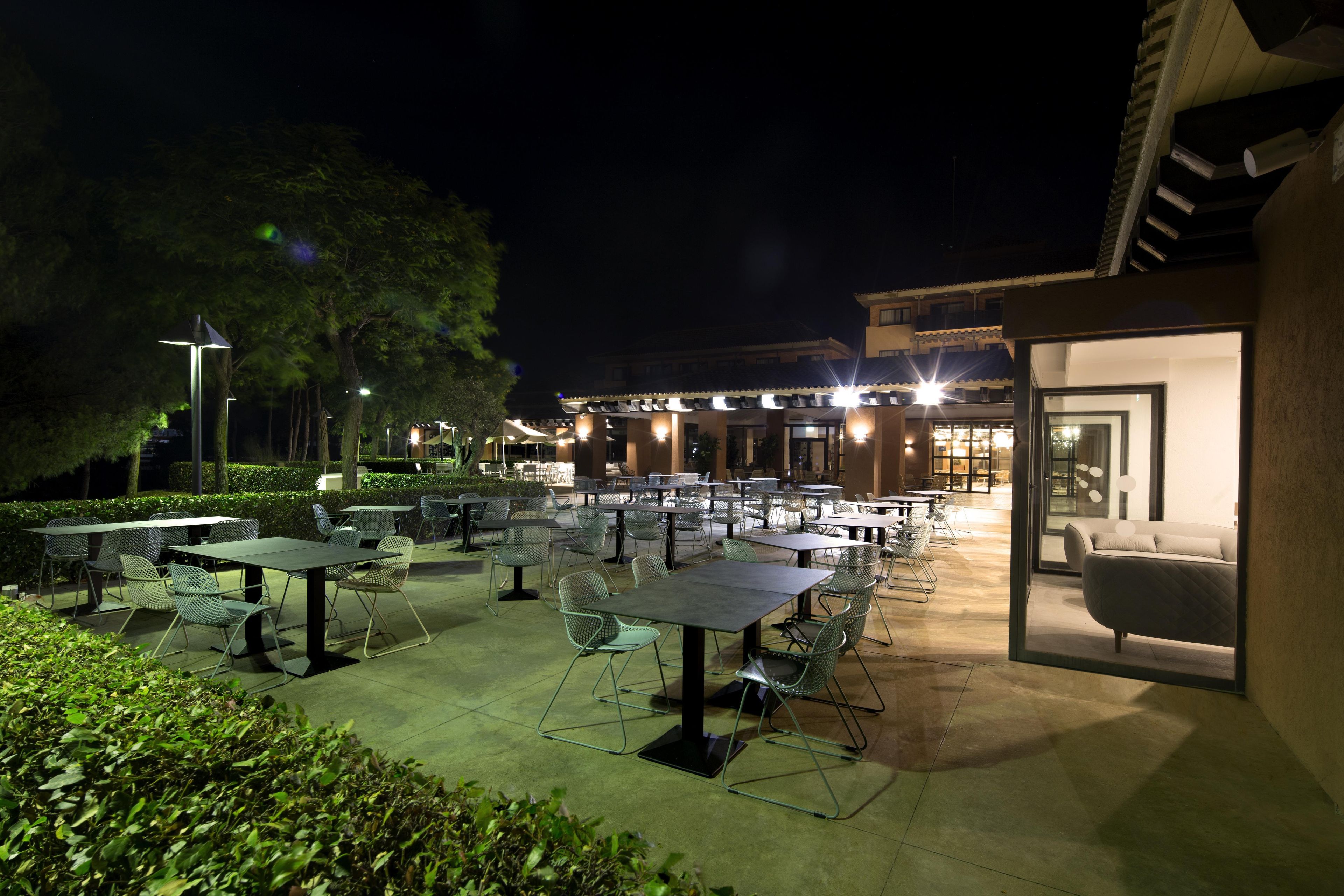 An outdoor dining area with tables and chairs, a building in the background, and a well-lit entrance.