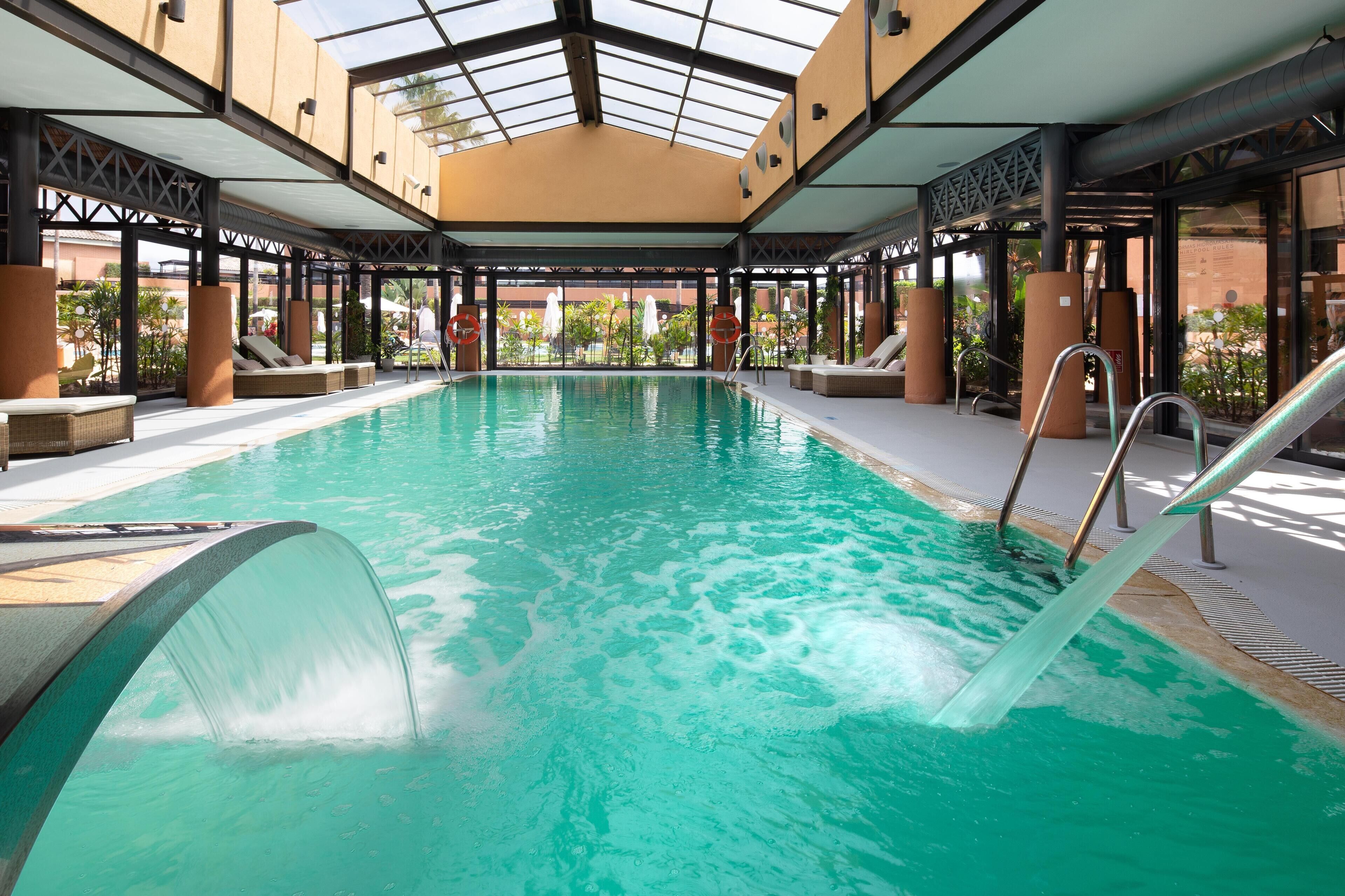 An indoor swimming pool with clear water, surrounded by lounge chairs and a covered area with a glass ceiling.