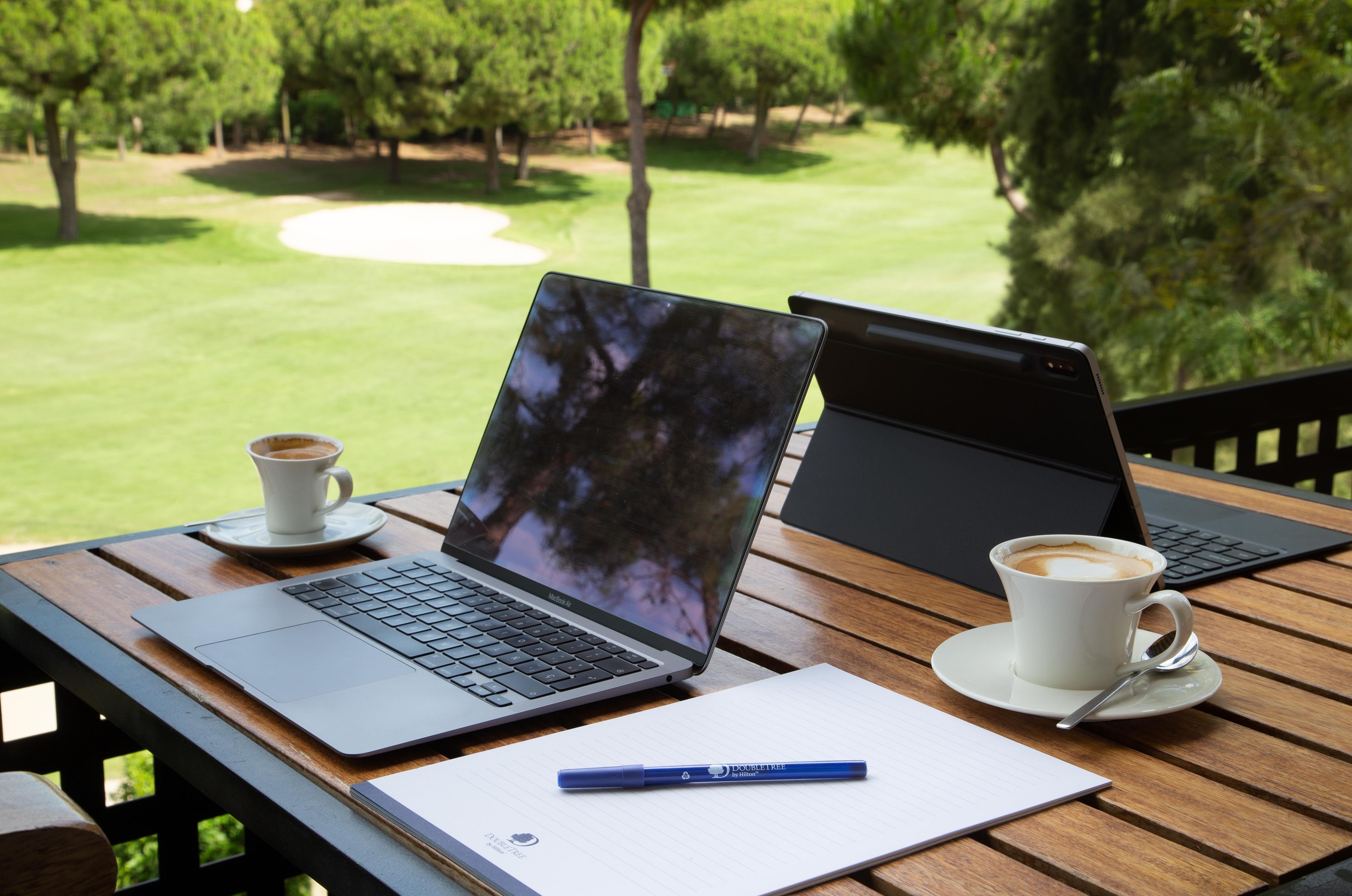 Two laptops on a wooden table with a cup of coffee and a notebook with a pen.