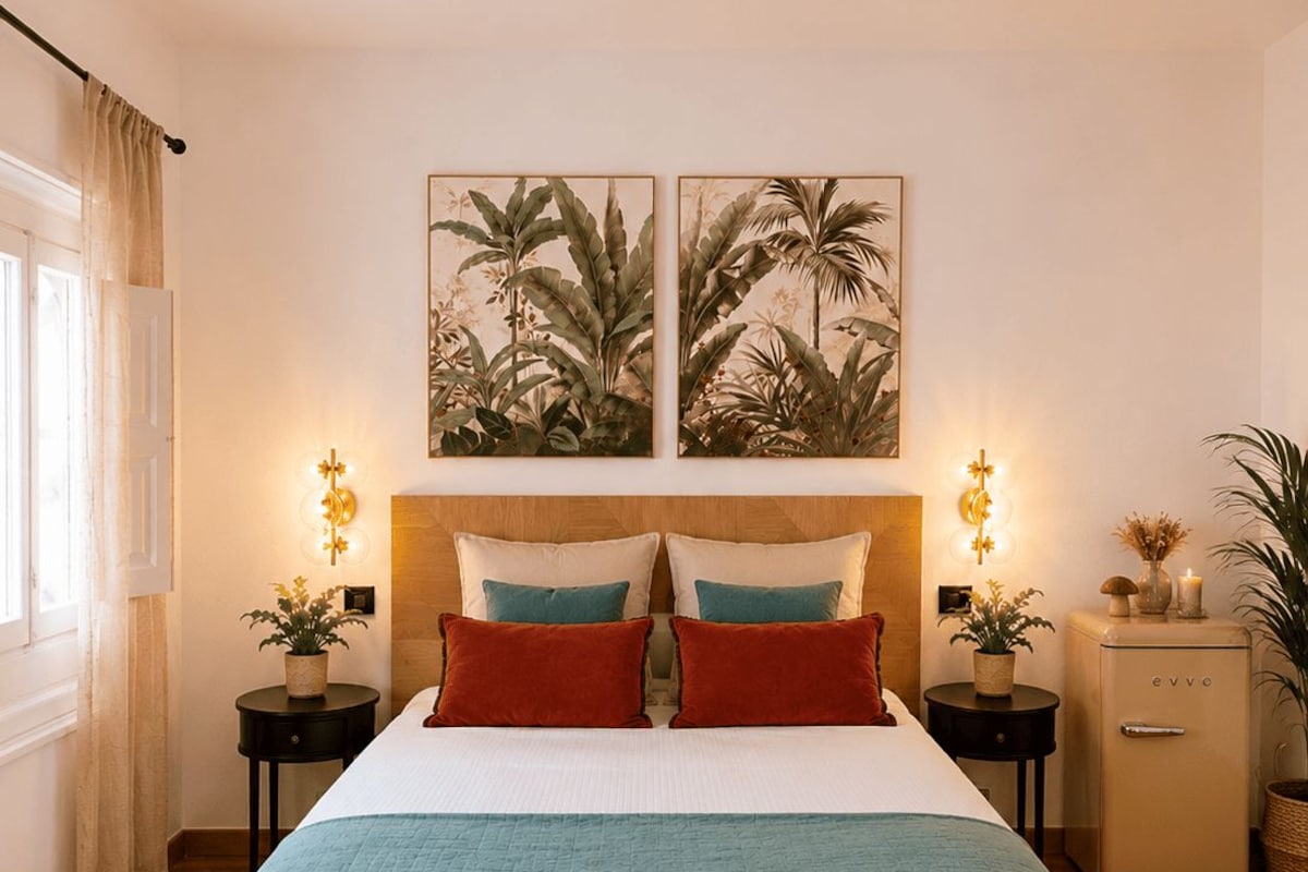 Una cama doble con colcha verde azulada y blanca, dos almohadas rojas y un cabecero de madera. Hay dos lámparas de pared y dos cuadros enmarcados con motivos botánicos sobre la cama.
