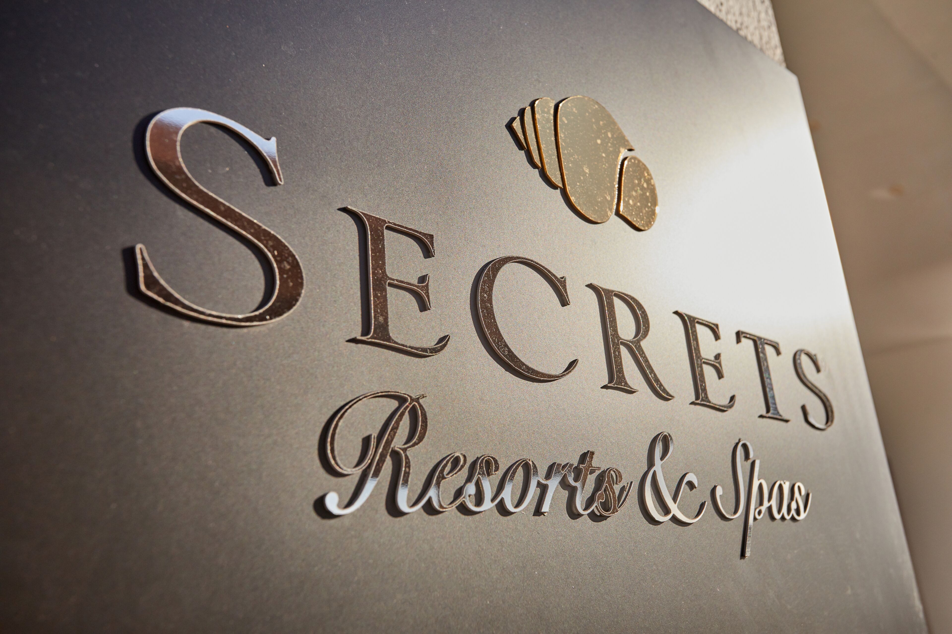 Ein Schild mit der Aufschrift "SECRETS Resorts & Spas" in metallischer Schrift, versehen mit einem dekorativen Element über dem Buchstaben 'E'.
