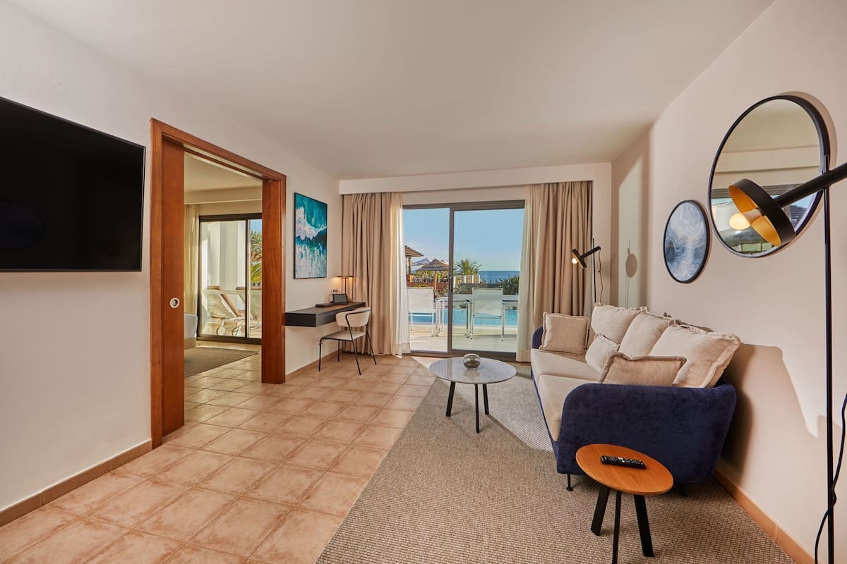 A hotel room with a sofa, a small table, a TV, a desk, and a view of the pool and beach.