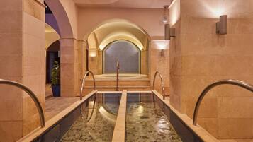 A modern indoor pool with clear water, surrounded by beige stone walls and a central archway leading to a seating area.