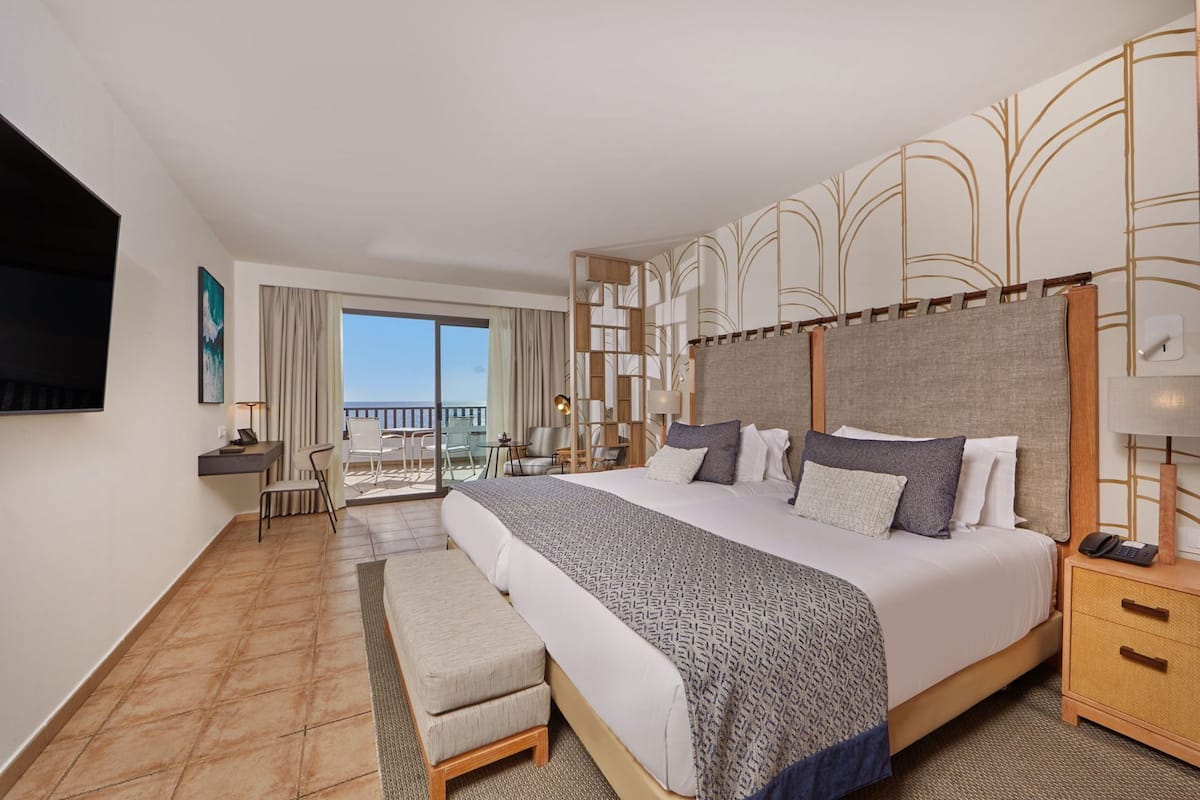 A hotel room with a large bed, a desk, a chair, a TV, and a balcony with a view.