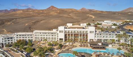Um resort com várias piscinas, um edifício central com varandas e um cenário montanhoso ao fundo.