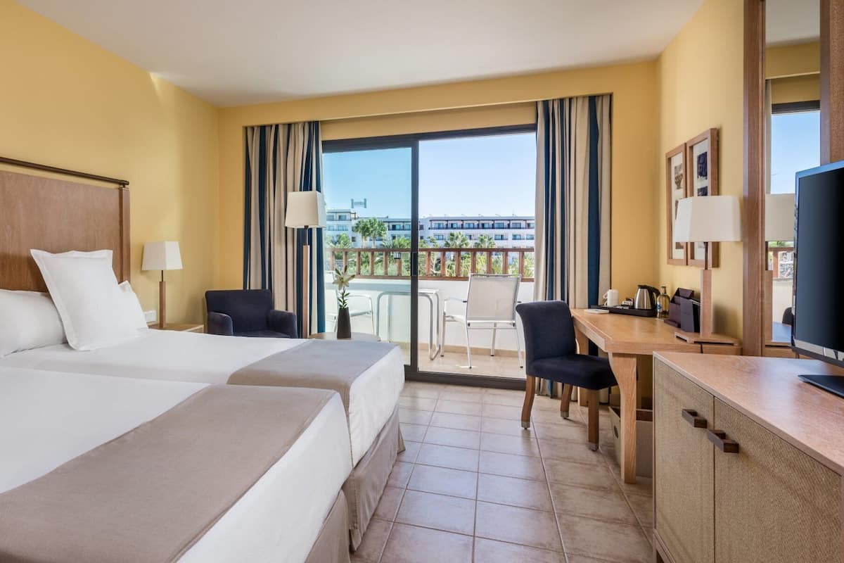 A hotel room with two beds, a desk, a TV, and a balcony with a view of buildings and palm trees.