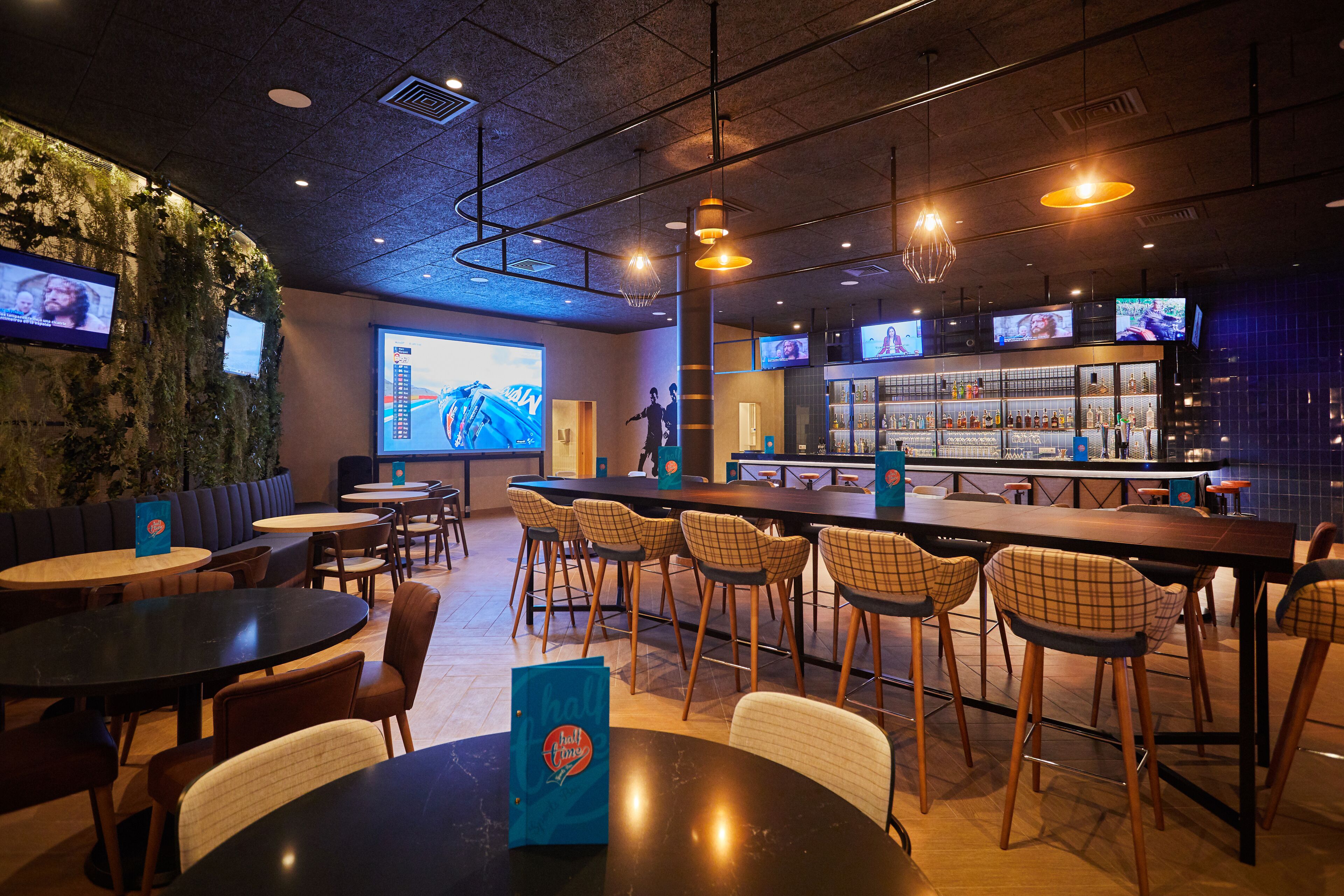A modern bar area with multiple seating options, including bar stools and tables, and a projection screen displaying a sports event.