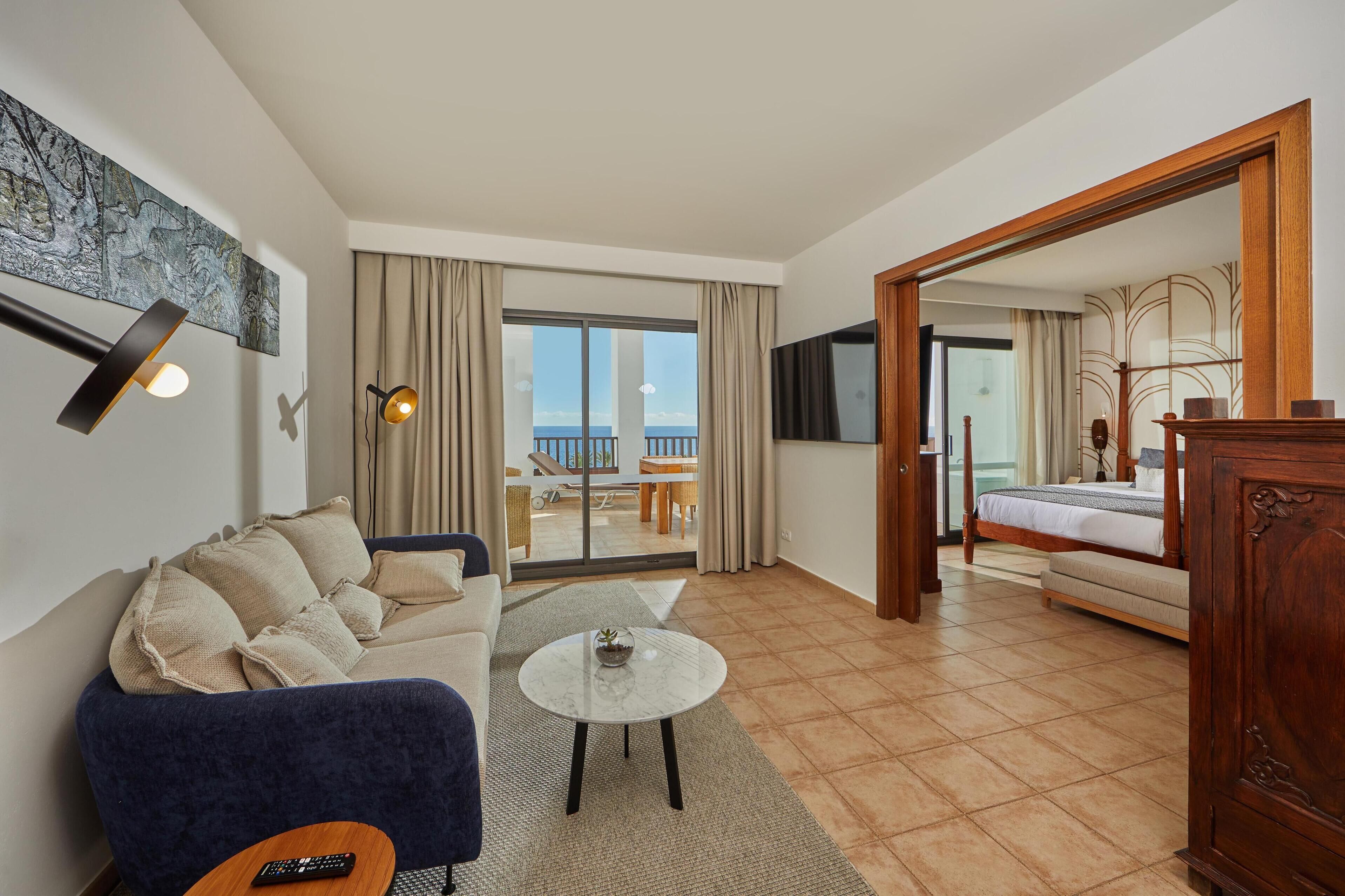 A modern hotel room with a sofa, a round coffee table, a bed, and a balcony with a view.