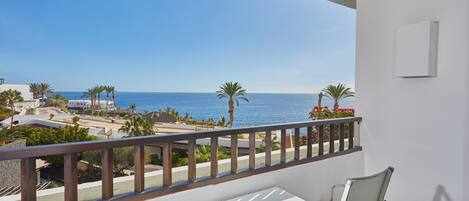 A balcony with a white table and chairs, overlooking a coastal area with palm trees and a clear blue sky.