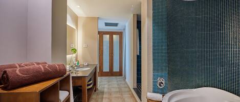 Un baño moderno con bañera, zona de ducha con azulejos azules, tocador de madera con toallas y una puerta que da a otra habitación.