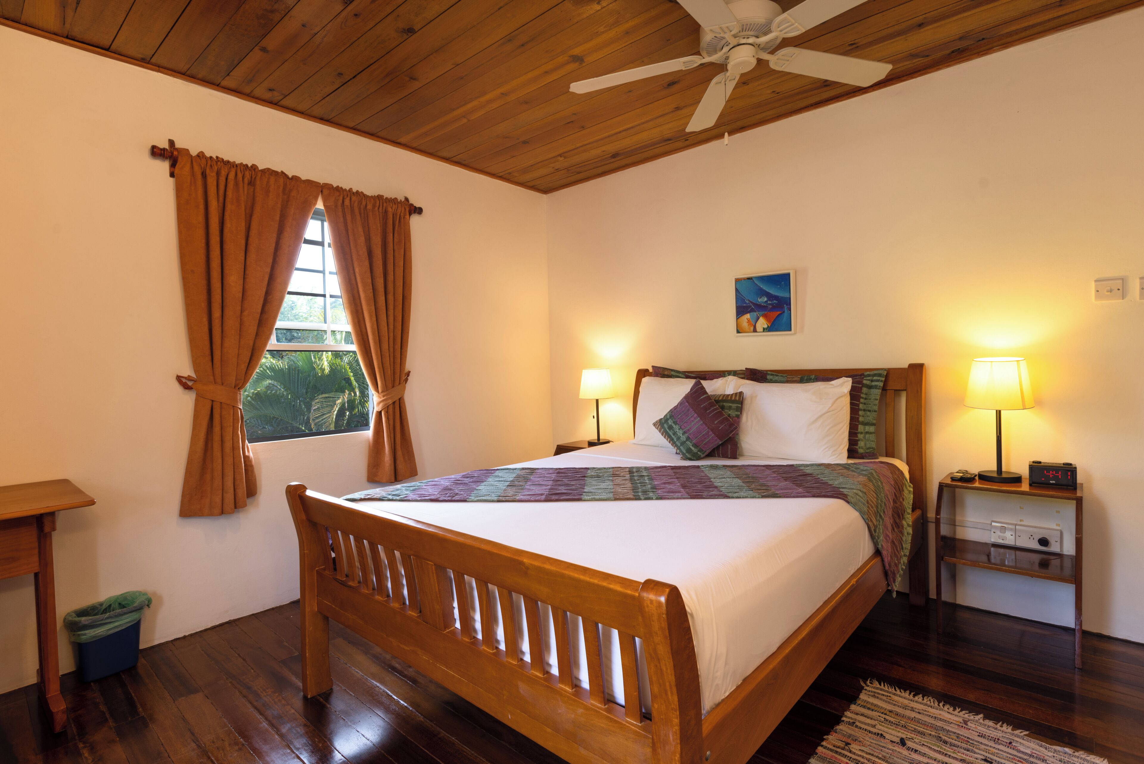 A bedroom with a wooden bed, two bedside lamps, a ceiling fan, and a window with curtains.