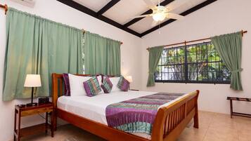A bedroom with a wooden bed, green curtains, a ceiling fan, and a window with a view of greenery.